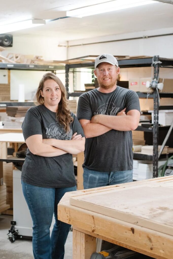 A photo of a man and a women, standing in a wood shop with their arms crossed and smiling at the camera.