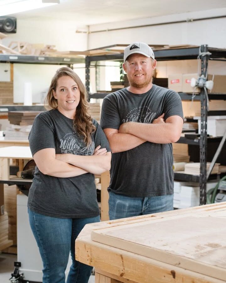 A photo of a man and a women, standing in a wood shop with their arms crossed and smiling at the camera.